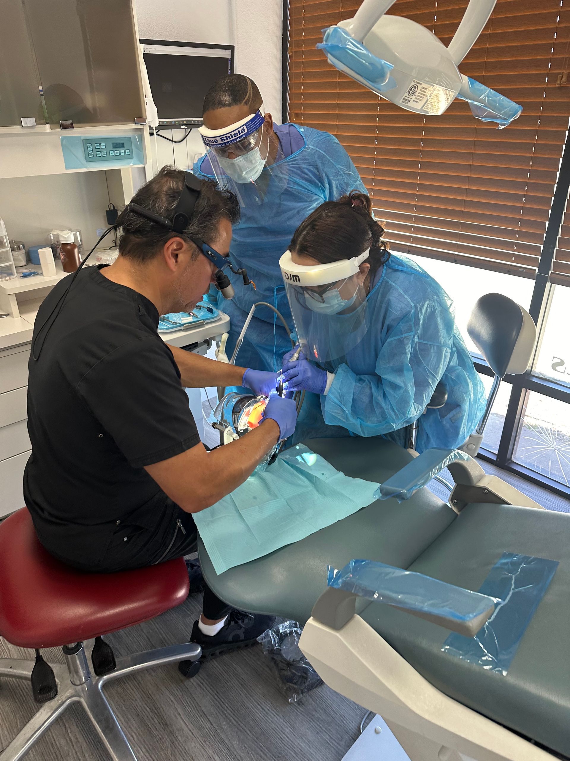 Dentist, assistant, and hygienist in a dental office, all wearing protective gear, working on a patient.