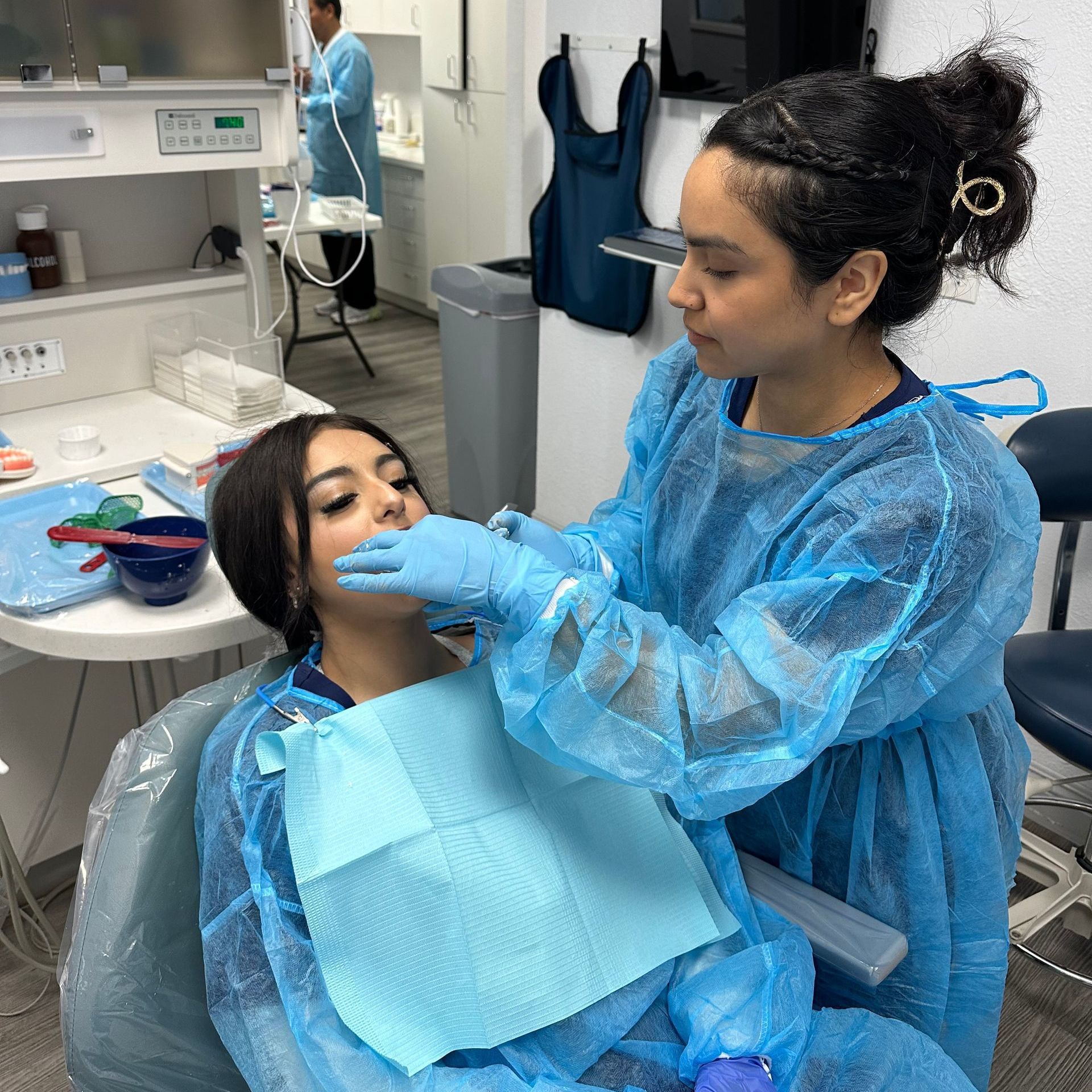 Dentist examining a patient's mouth in a dental office. Both wear blue protective clothing and gloves.