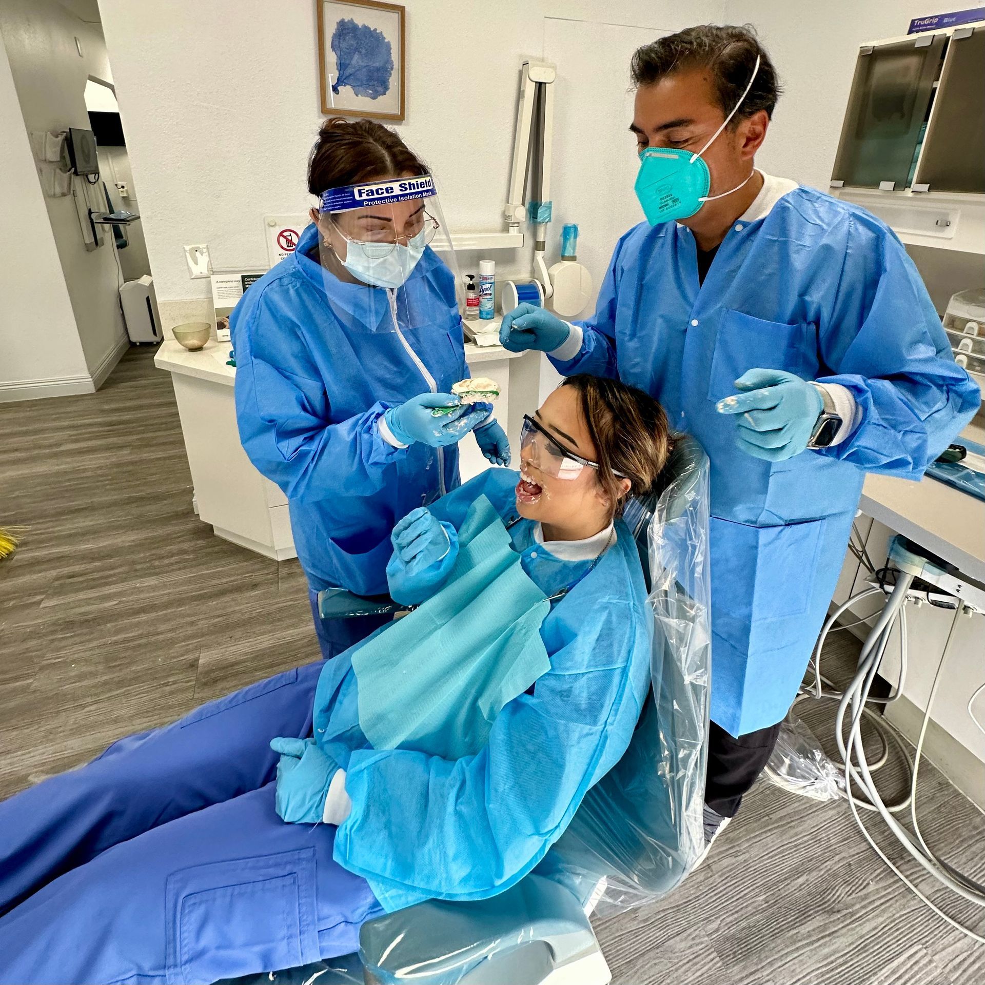 Dentists in blue protective gear examine a patient's teeth in a dental office.