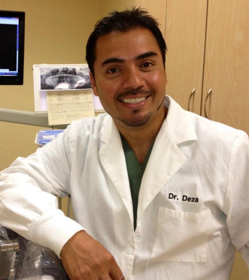Smiling Dr. Deza in a white coat, leans on dental equipment, inside a clinic.