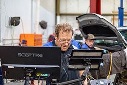 Mechanic using computer at auto repair shop; another works on a car with the hood open.