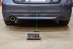 A car's rear bumper with a laser alignment tool on the ground projecting a vertical green line.