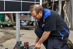 Mechanic kneels, examining equipment in a shop. A black grid is behind him.