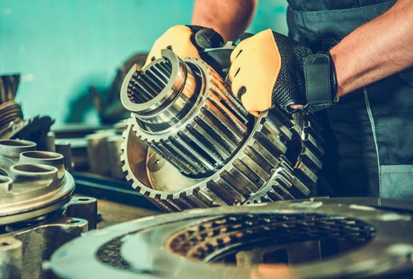 Mechanic holding a gear assembly, wearing yellow gloves, in a workshop.