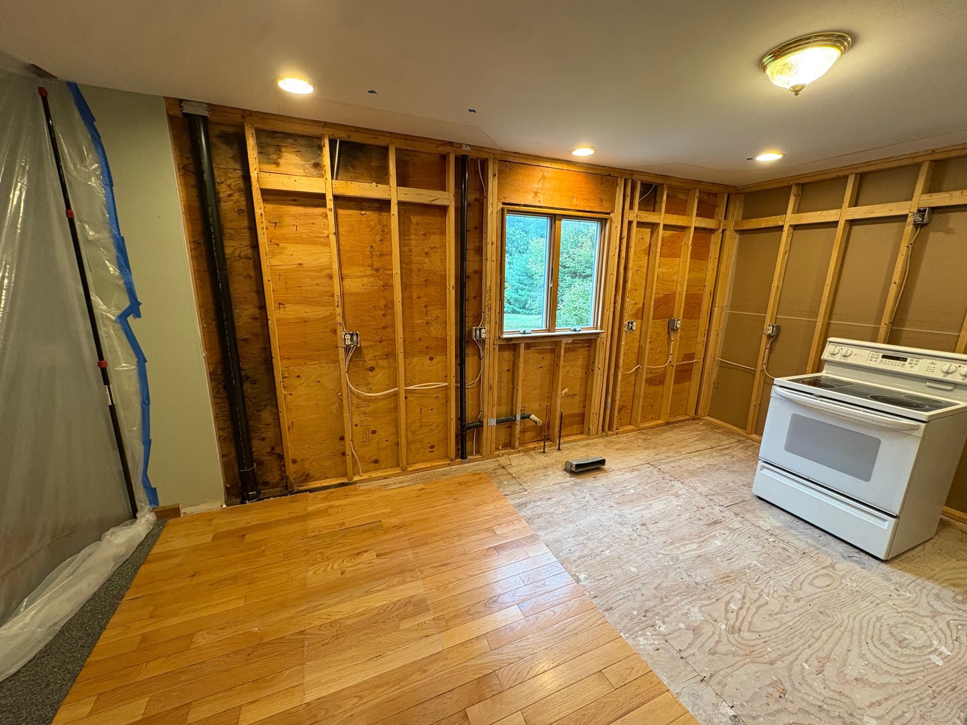 A room under renovation with exposed wood wall studs, a window, hardwood flooring, and a white oven.
