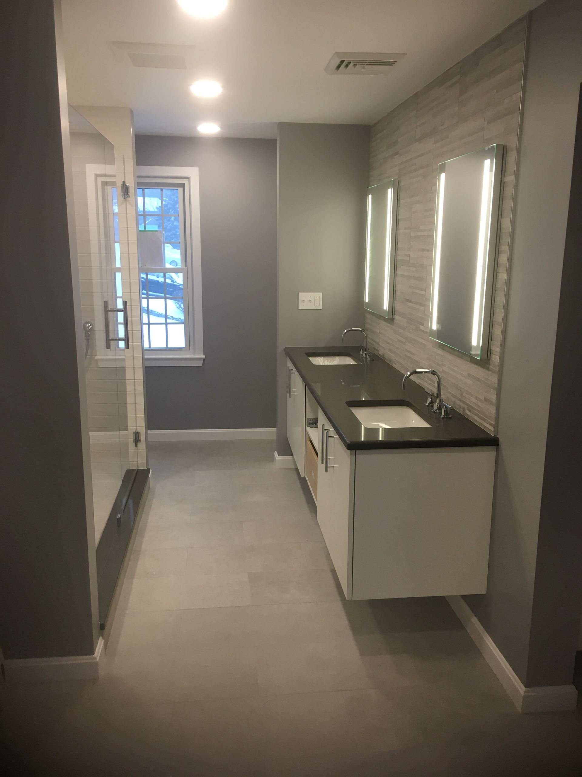 Modern bathroom with gray tile, double vanity, mirrors, and a shower in the hallway view