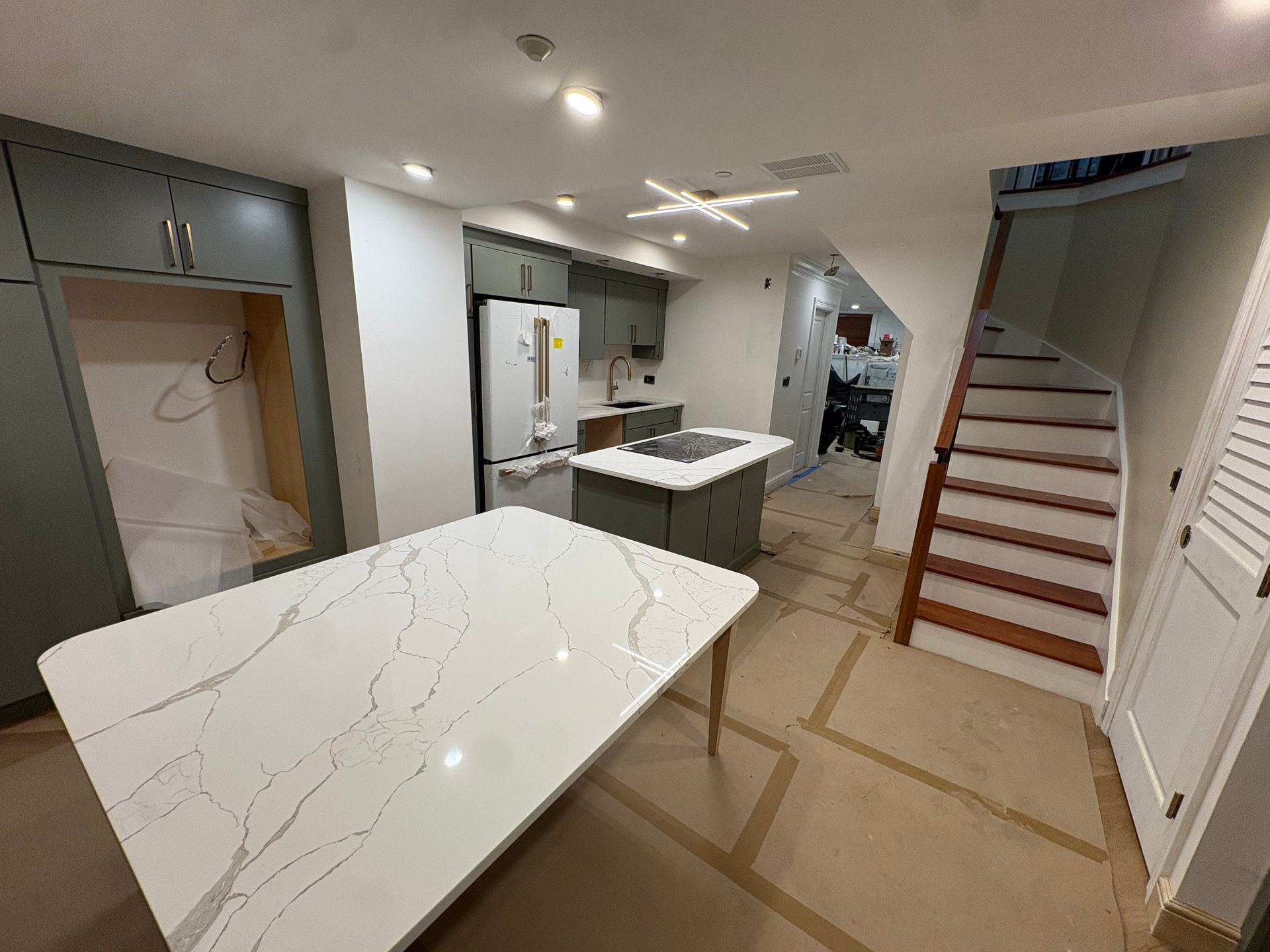 A modern kitchen undergoing renovation with white stone-topped islands, grey cabinets, and wooden stairs.