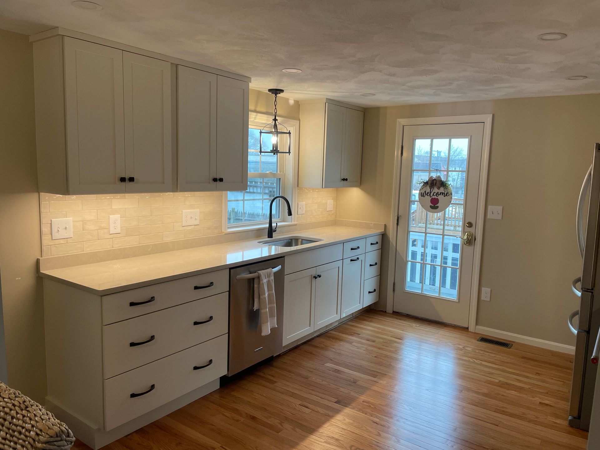 A kitchen featuring light-colored cabinets, a stainless steel dishwasher, a window over the sink, and wood flooring.