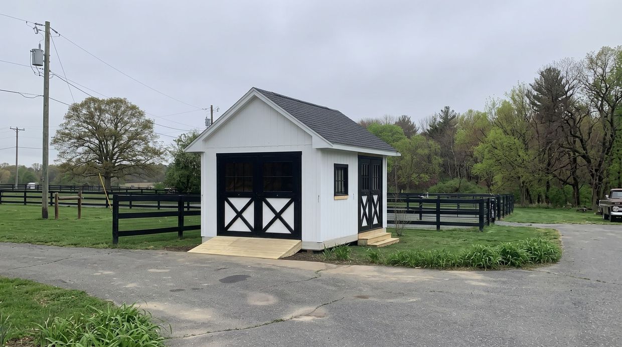 Small white barn-style shed with black trim beside a fenced rural driveway under cloudy skies