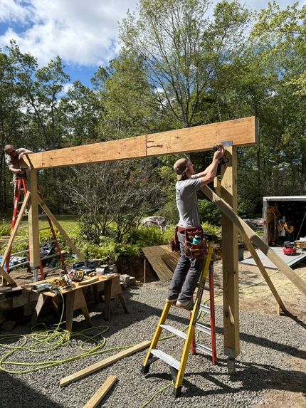 Two people lifting a long wooden beam outdoors, building a frame on a sunny gravel site.