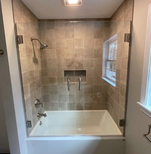 Bathroom with a white bathtub, glass shower doors, tan tiled walls, and a window.