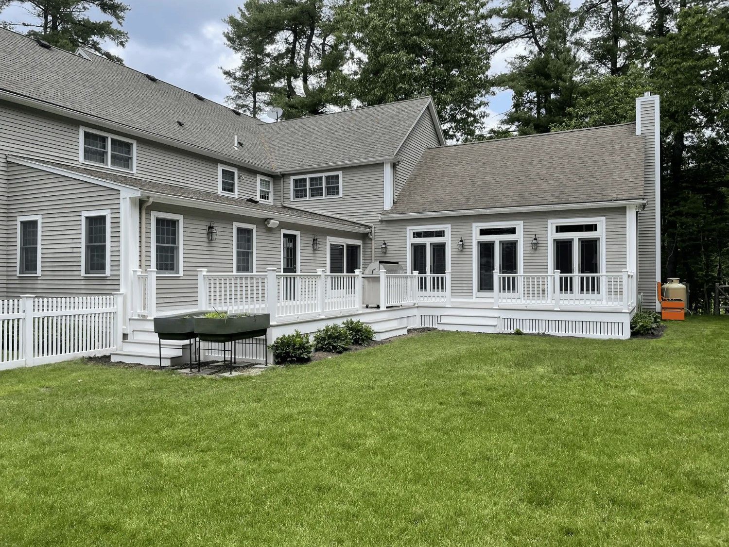 Backyard of a house with a deck, pergola, and green lawn. Gray siding, white railings, and an orange propane tank.