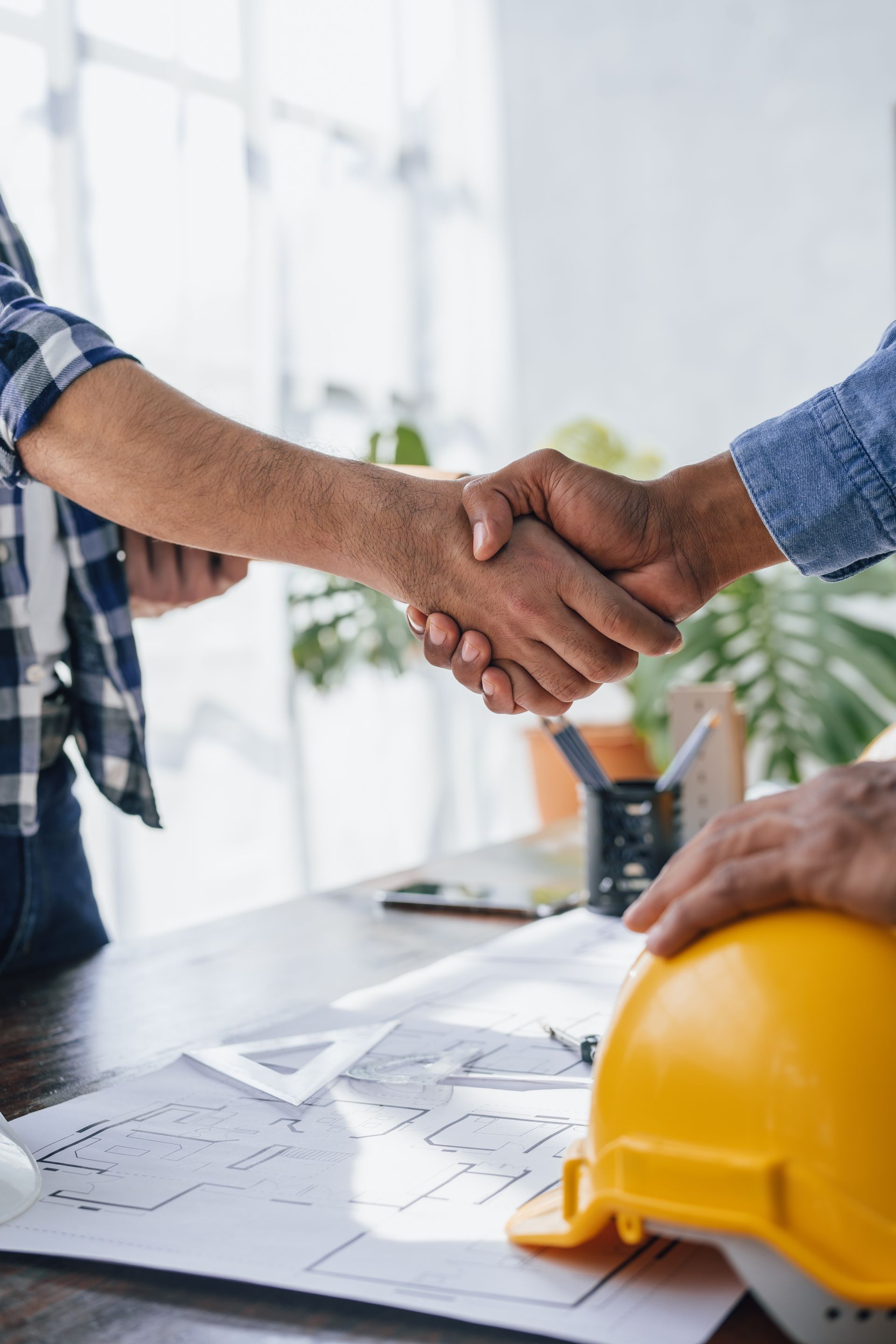 Two men are shaking hands over a table with a hard hat on it.
