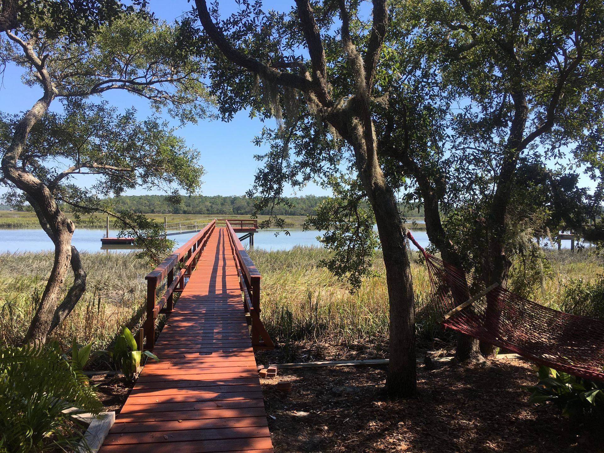 Wooden dock extends over marshy water towards a structure in the distance, under a blue sky.