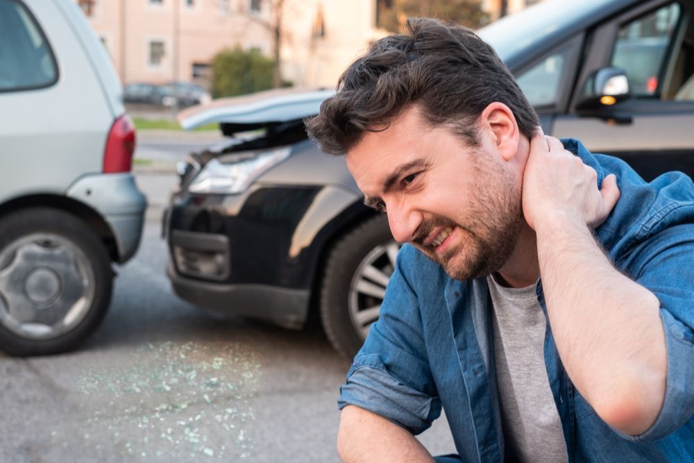 Man holding neck in pain after a car accident; damaged cars on street.