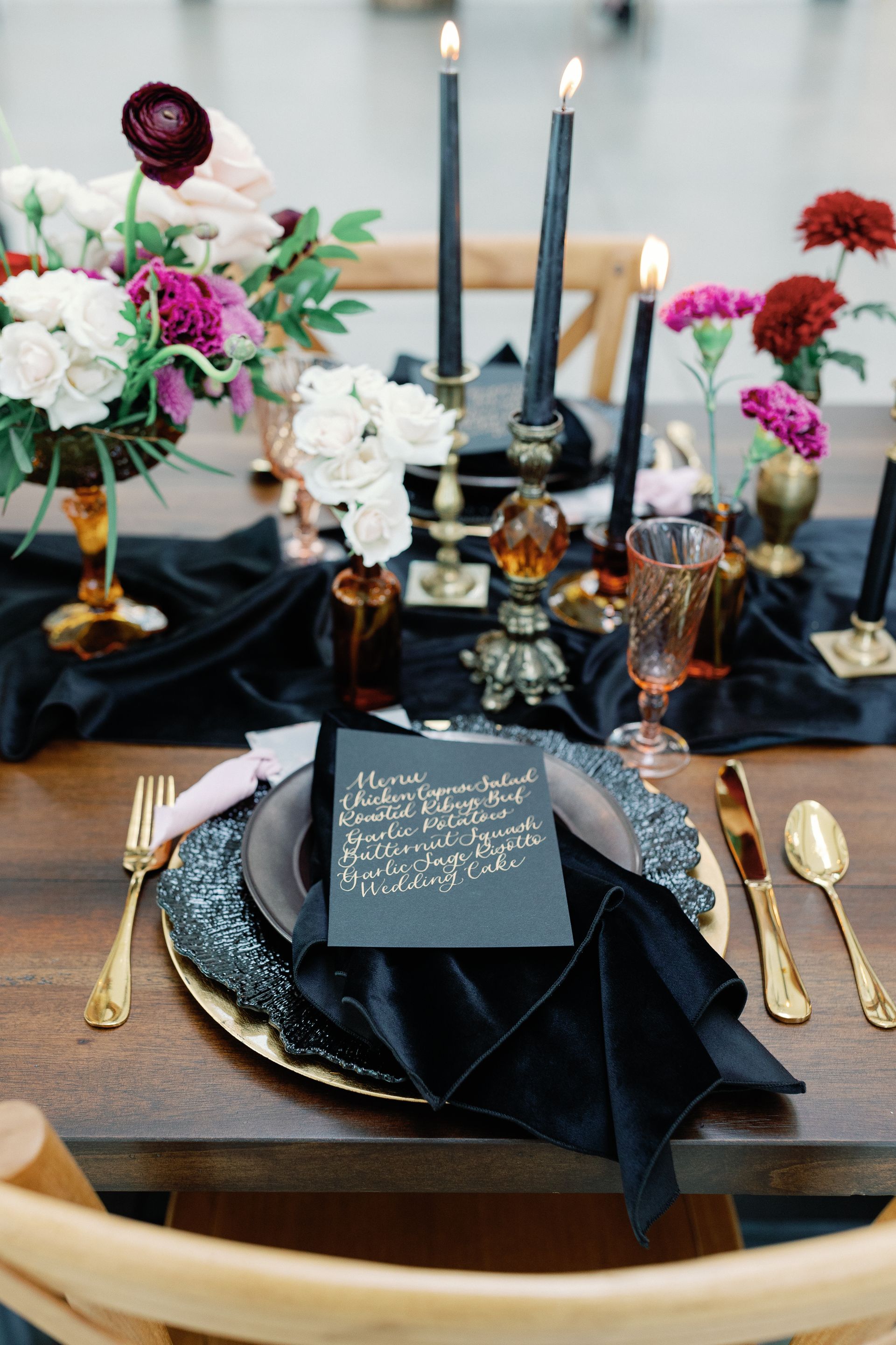 A table setting with black plates , black candles , gold silverware and flowers.