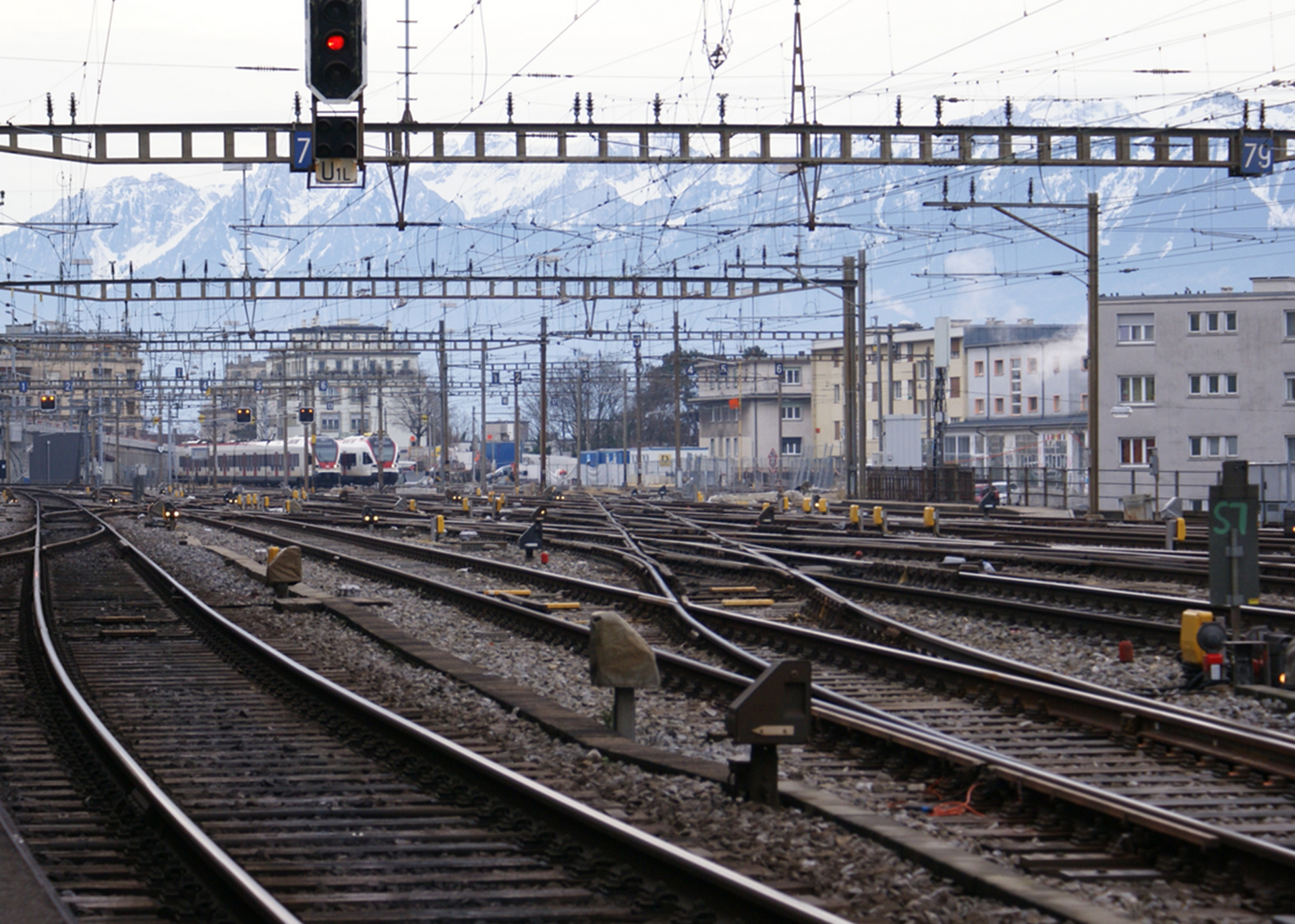 Plusieurs voies ferrées convergent à proximité de la gare de Lausanne, avec des lignes électriques aériennes et des montagnes enneigées à l'horizon.