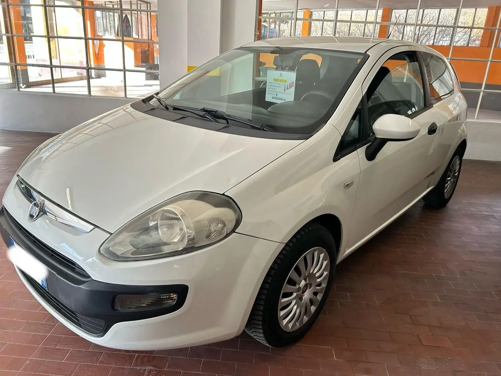 Un'auto hatchback bianca parcheggiata all'interno su un pavimento piastrellato vicino a grandi finestre.