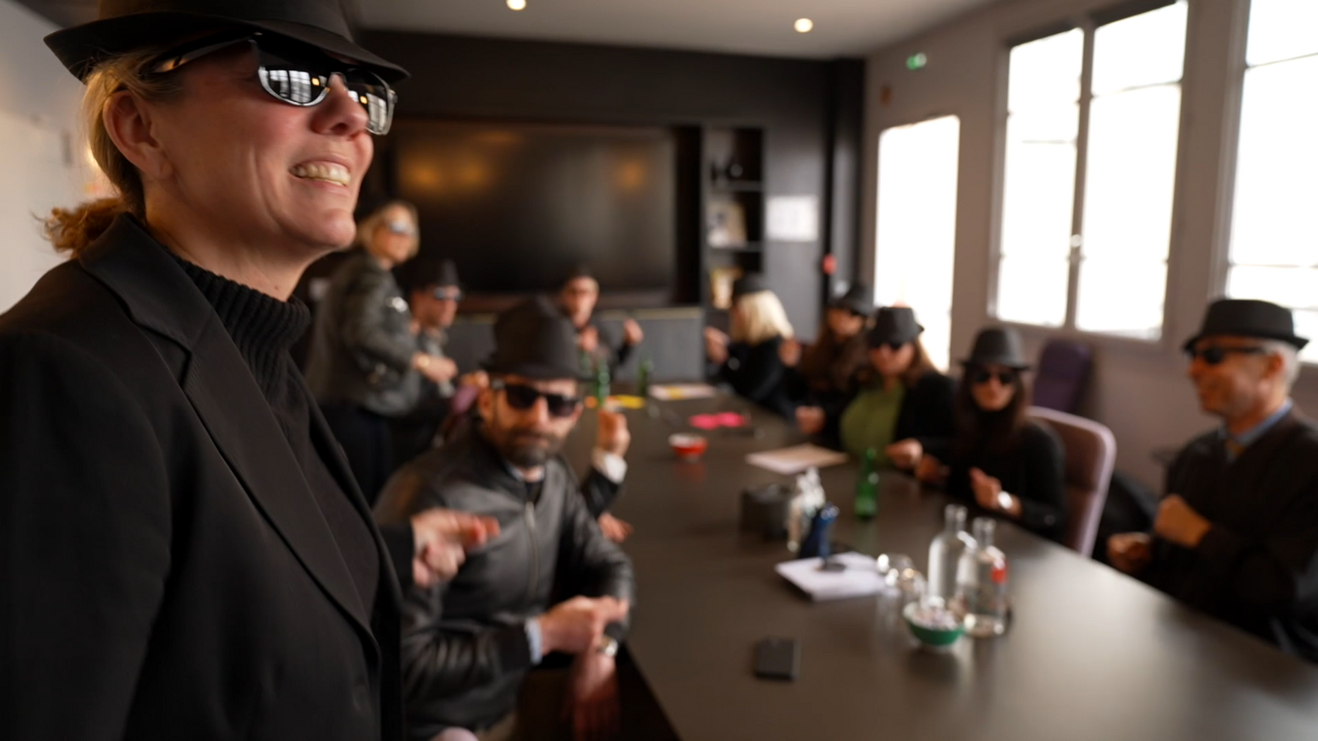 Une femme en costume noir et chapeau se tient devant un groupe de personnes assises autour d'une table.