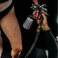 Two people in black clothing, one holding a metallic spray gun with a hose against a dark background.