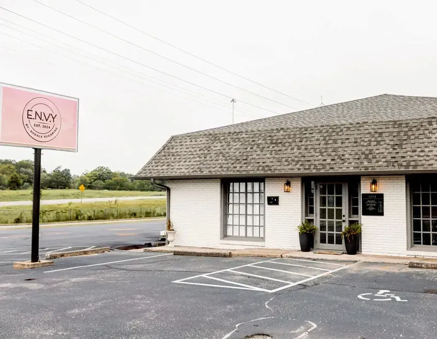Small storefront with pink sign, white exterior, and parking lot in front under a cloudy sky.