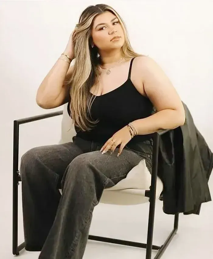 Woman seated on a chair, wearing a black top and gray pants, posing against a light background.