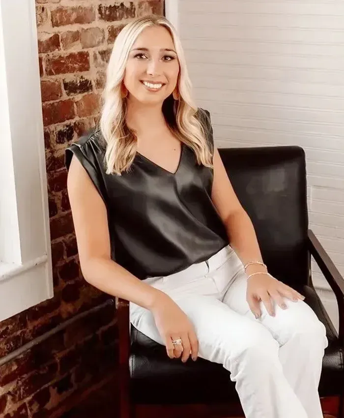 Smiling woman seated in a black chair beside a brick wall and window, wearing a black top and white pants