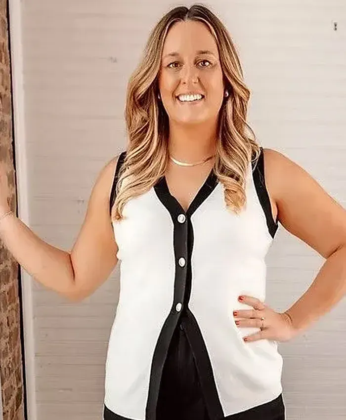 Smiling woman in a white and black sleeveless top standing indoors with one hand on her hip