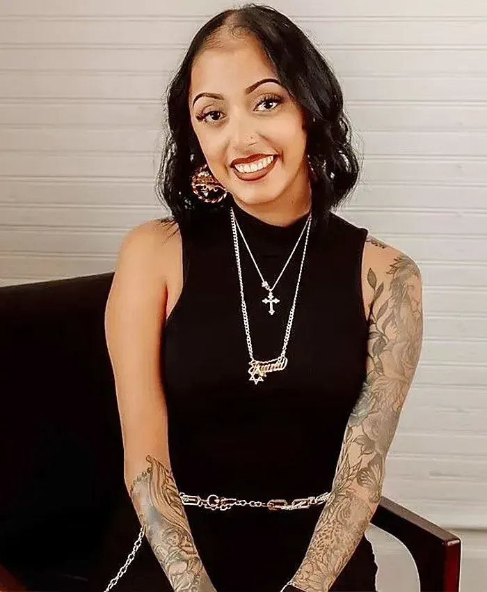 Smiling person seated in a black chair, wearing a black sleeveless top and layered necklaces, with tattooed arms.
