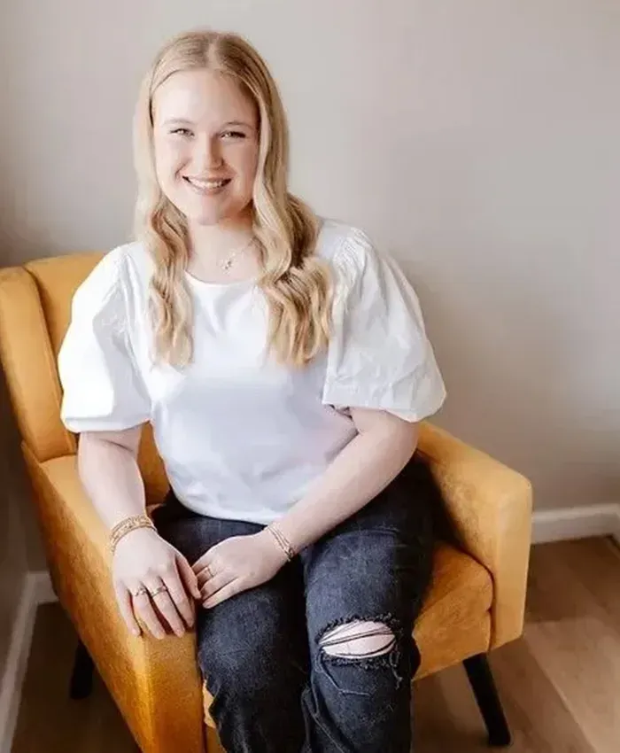 Person sitting in a mustard-yellow chair, smiling in a white blouse and black jeans in a bright room