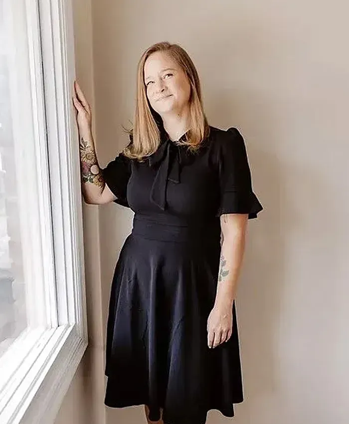 Woman in a black dress standing by a bright window, resting one hand on the curtain.
