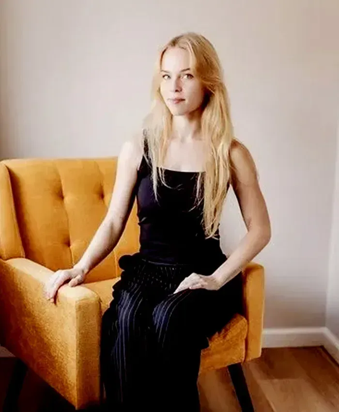 Woman sitting on a mustard armchair in a bright room, wearing a black dress and looking at the camera.