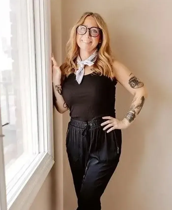 Woman in black outfit posing by a window, wearing glasses and a neck scarf.