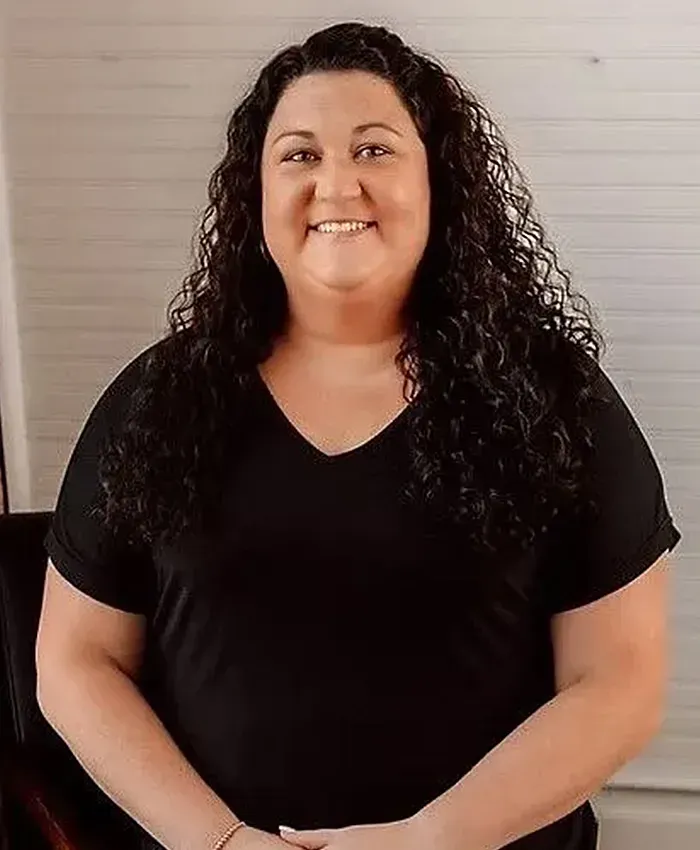 Smiling person in a black shirt standing indoors against a light wall.