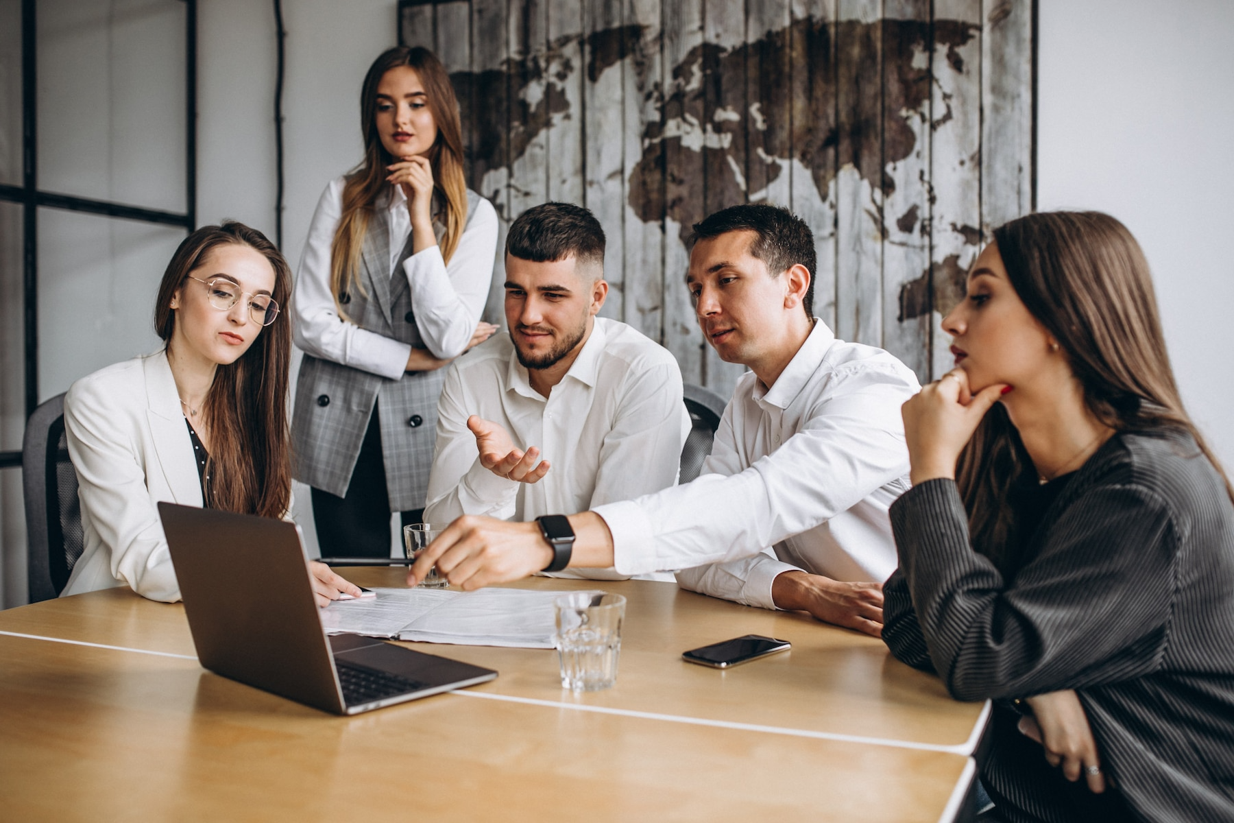 Five people in a meeting, focused on a laptop and papers on a wooden table. A world map is in the background.