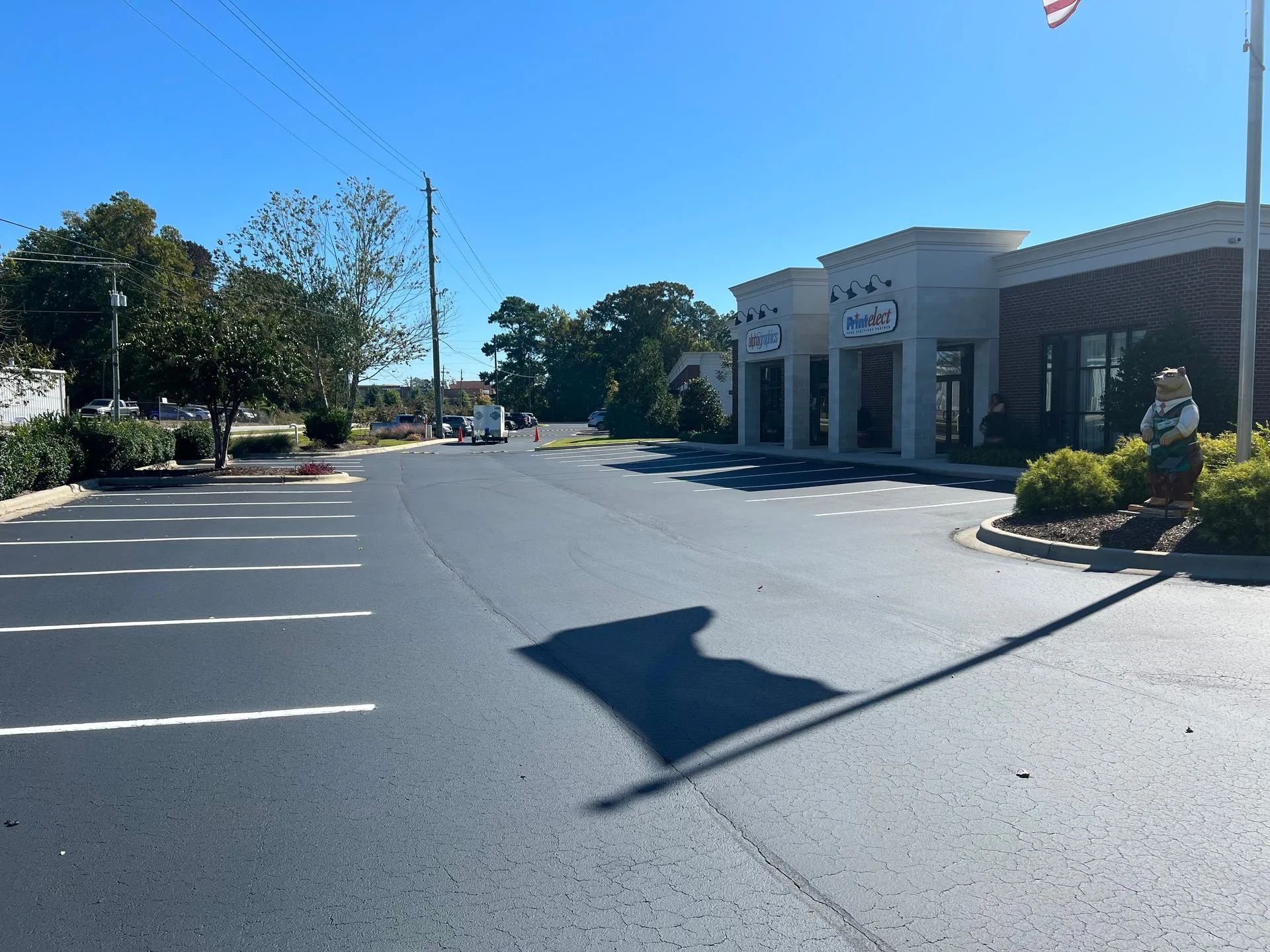 Empty parking lot beside a white commercial building on a sunny day, with trees and a flagpole in view