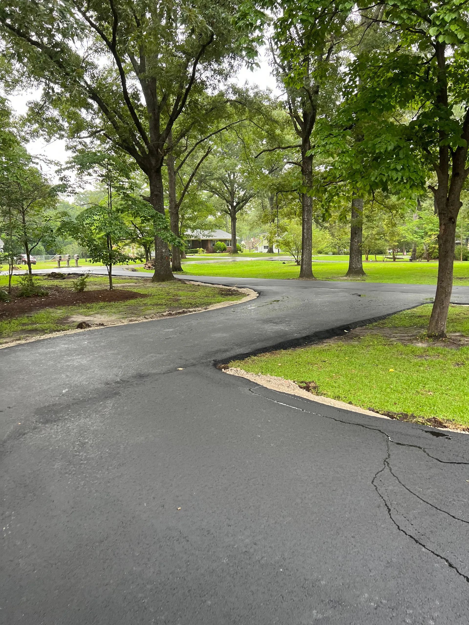 Black asphalt driveway curves through a park-like setting with trees and green grass.