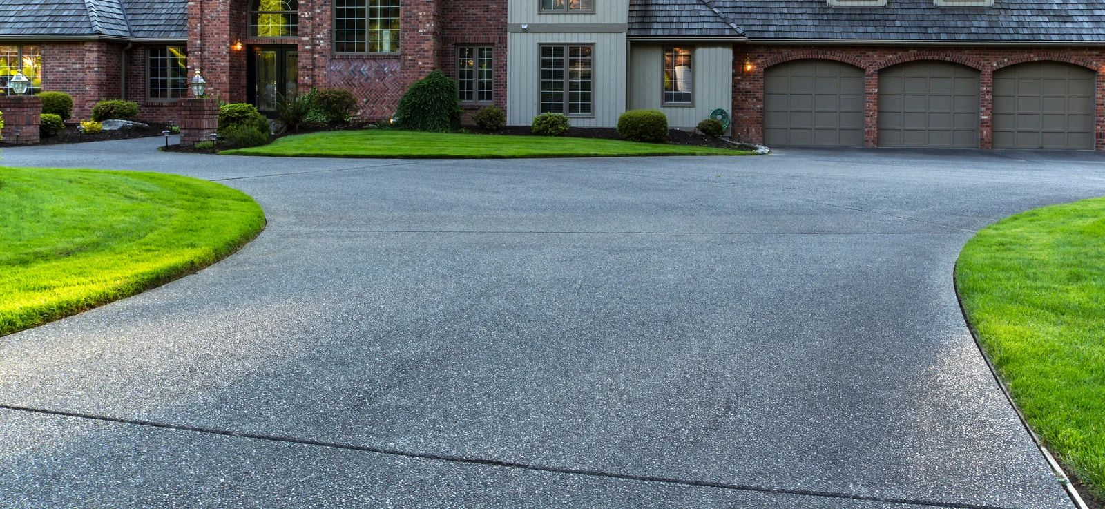 Curved asphalt driveway leading to a brick house with a three-car garage and green lawns