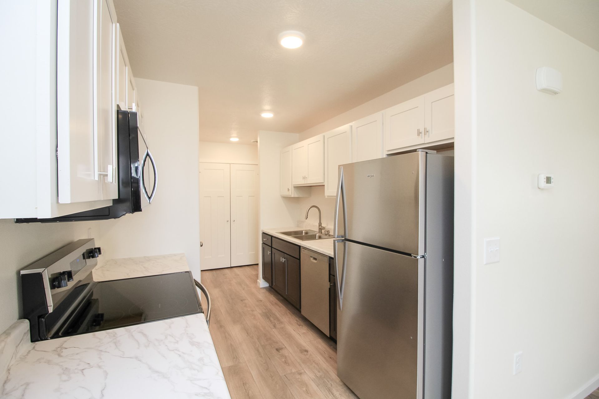 Narrow modern kitchen with stainless steel appliances, white cabinets, and light wood floors.