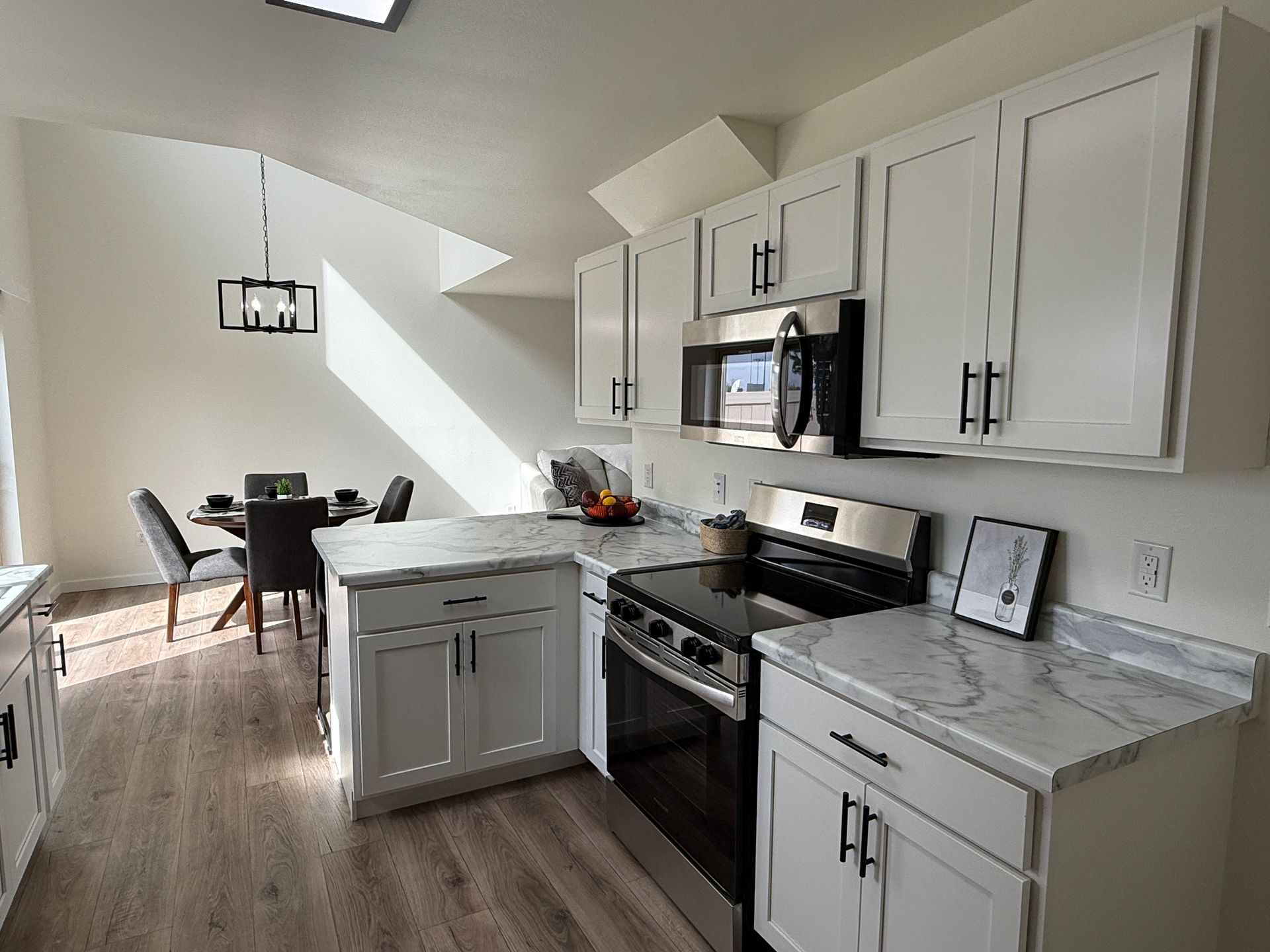 Bright kitchen with white cabinets, stainless appliances, gray counters, and a small dining table by a window.