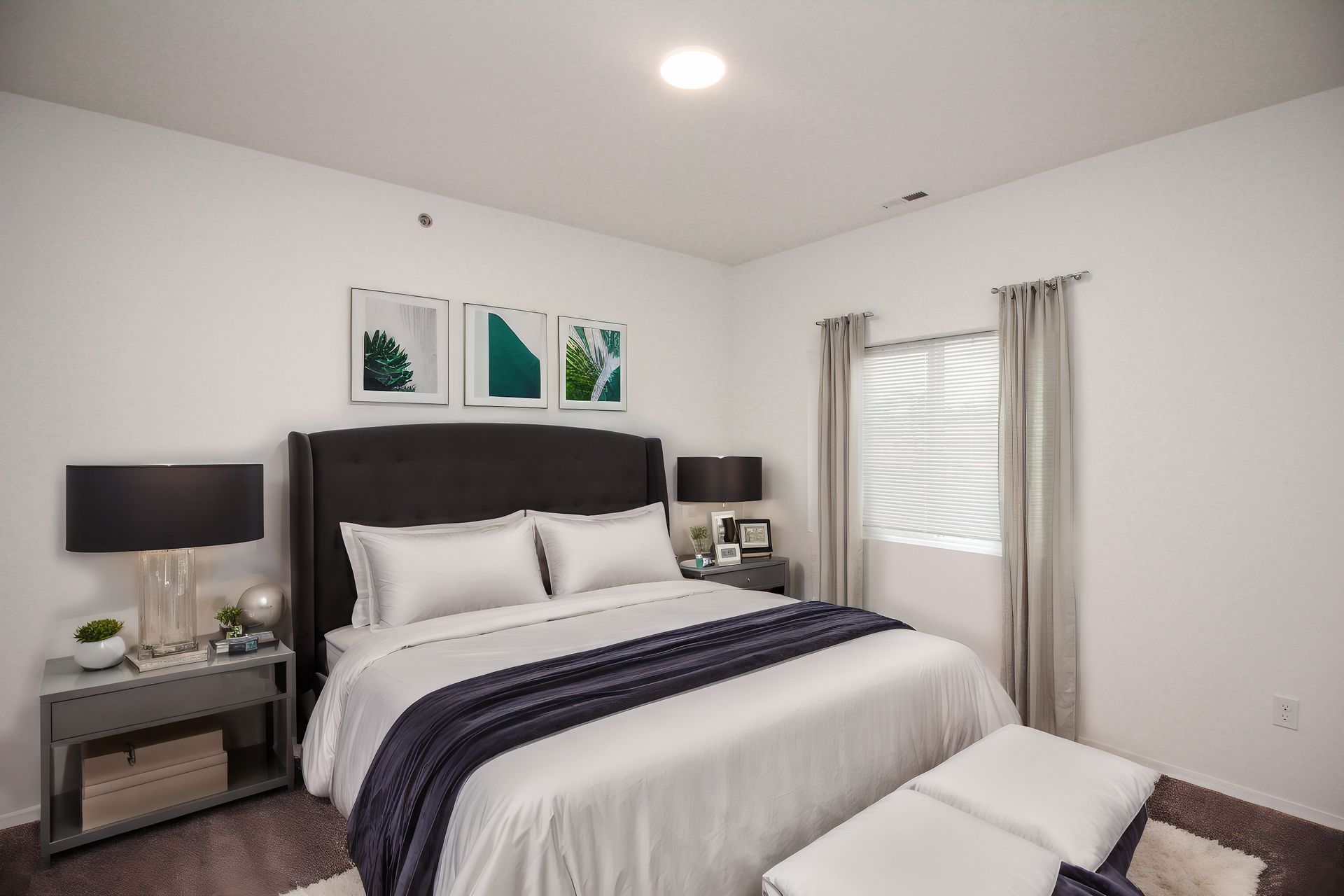 Bright modern bedroom with a dark upholstered bed, white bedding, nightstands, and a window with blinds and curtains