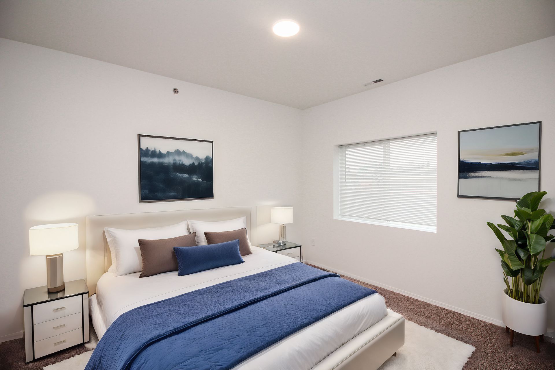 Bright bedroom with a blue bedspread, white lamps, wall art, and a window with blinds.