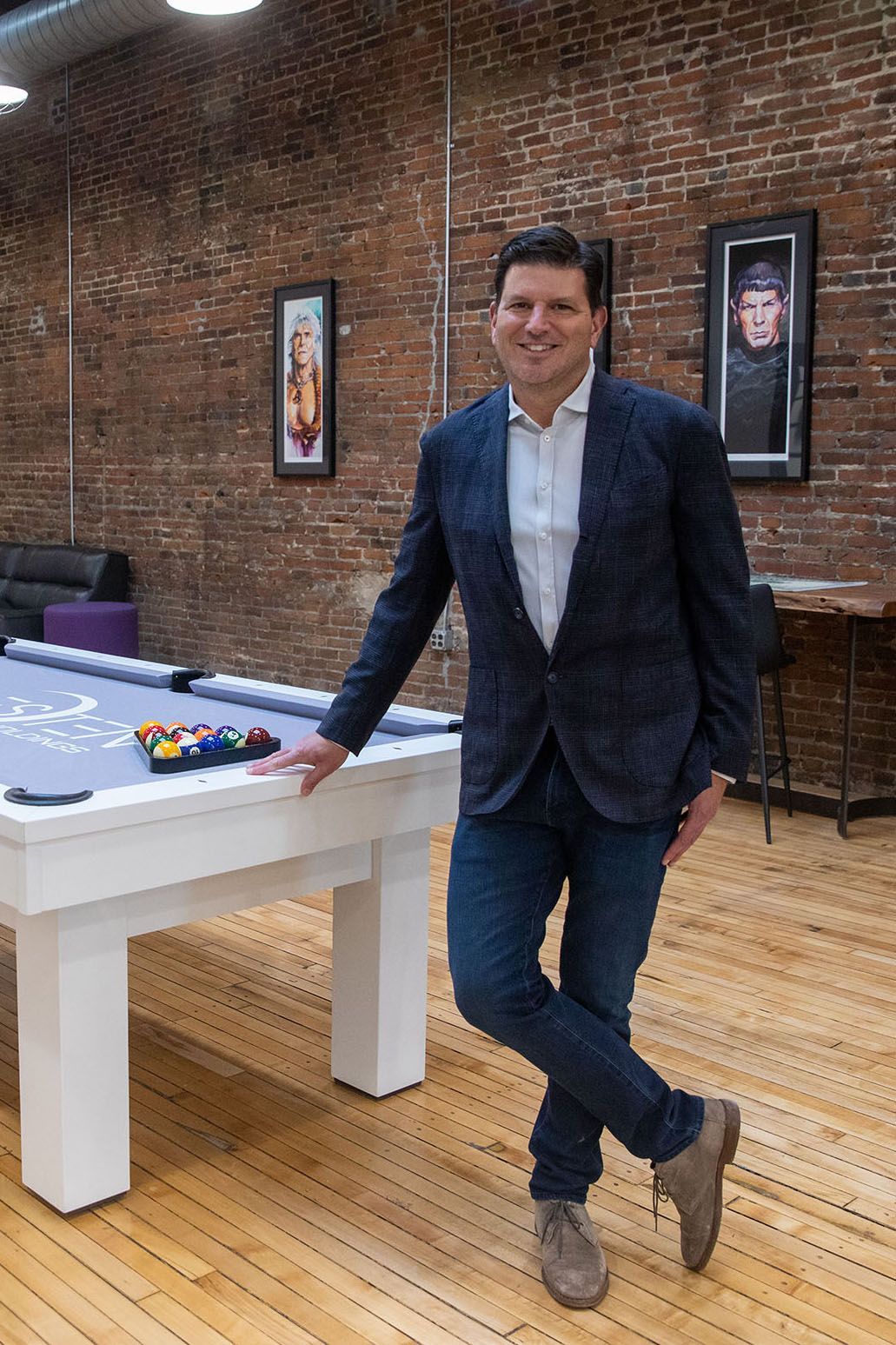 Man leaning on a white pool table in a brick-walled room with framed photos.
