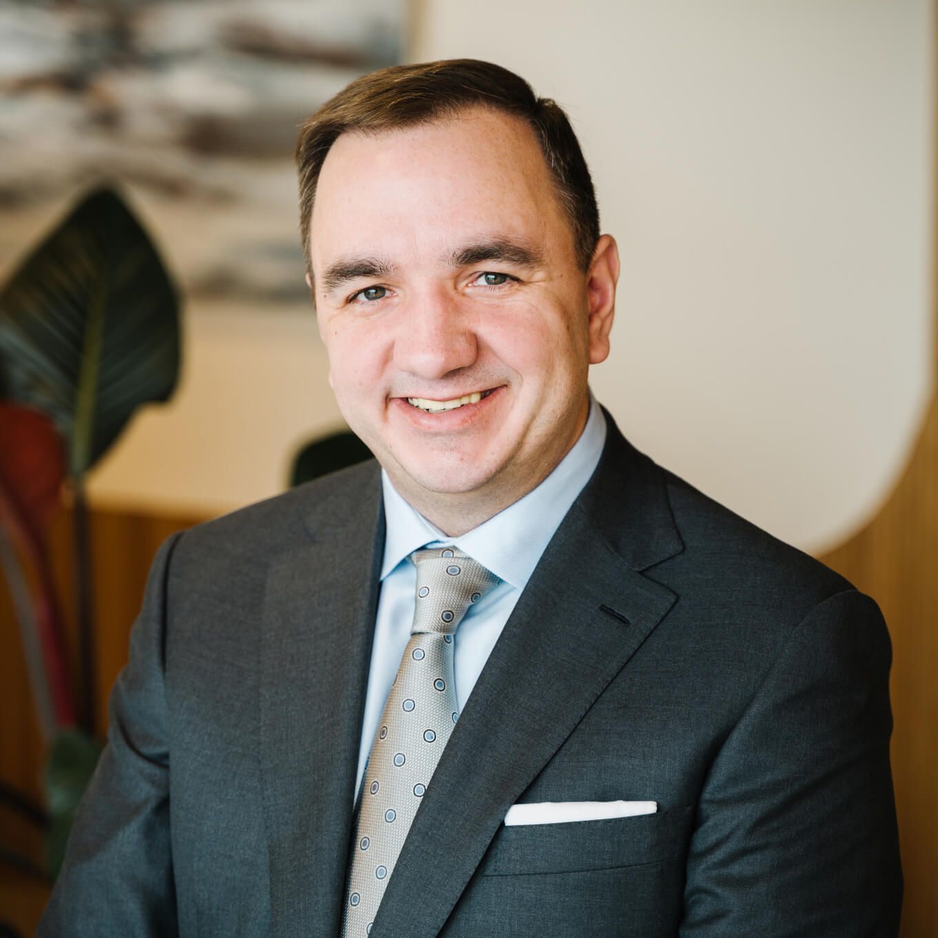 Smiling man in a dark suit and tie posing indoors against a blurred background