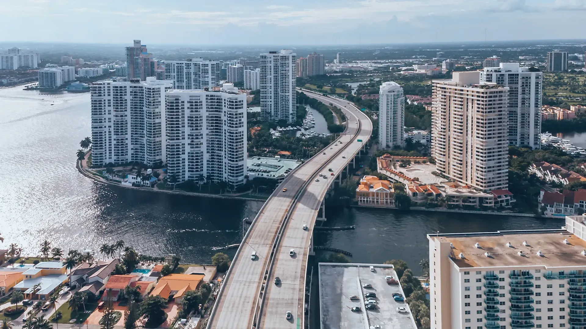 An elevated highway curves between high-rise apartment buildings along a waterfront in a sunny, developed coastal city.