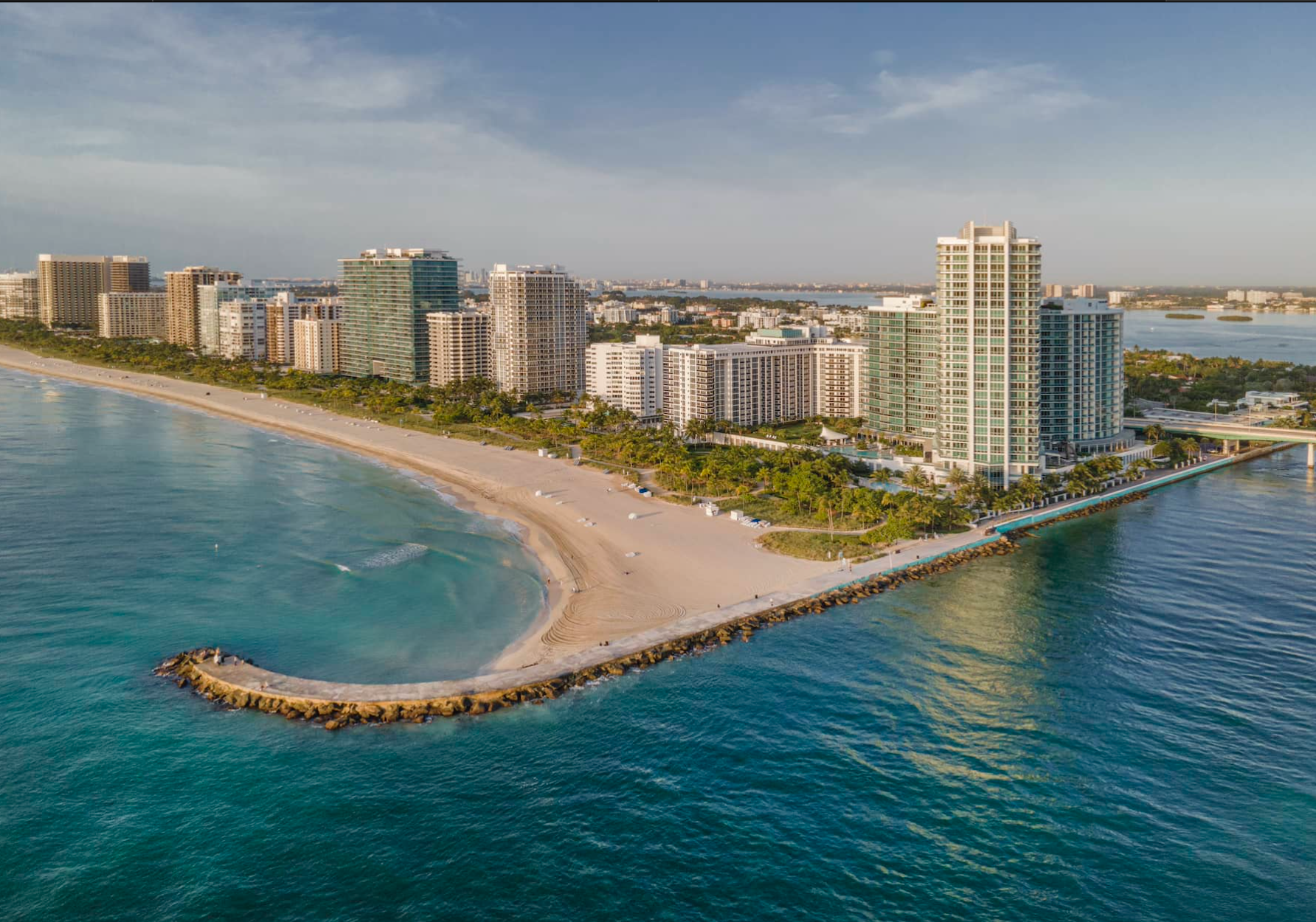 An aerial view of a sandy beach curving around a stone jetty, lined with high-rise apartment buildings and green trees.