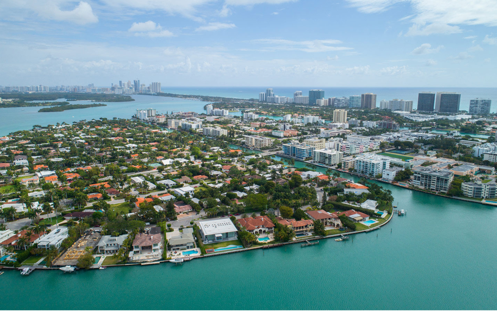 Aerial view of a coastal neighborhood with houses, lush trees, and swimming pools, surrounded by turquoise water.