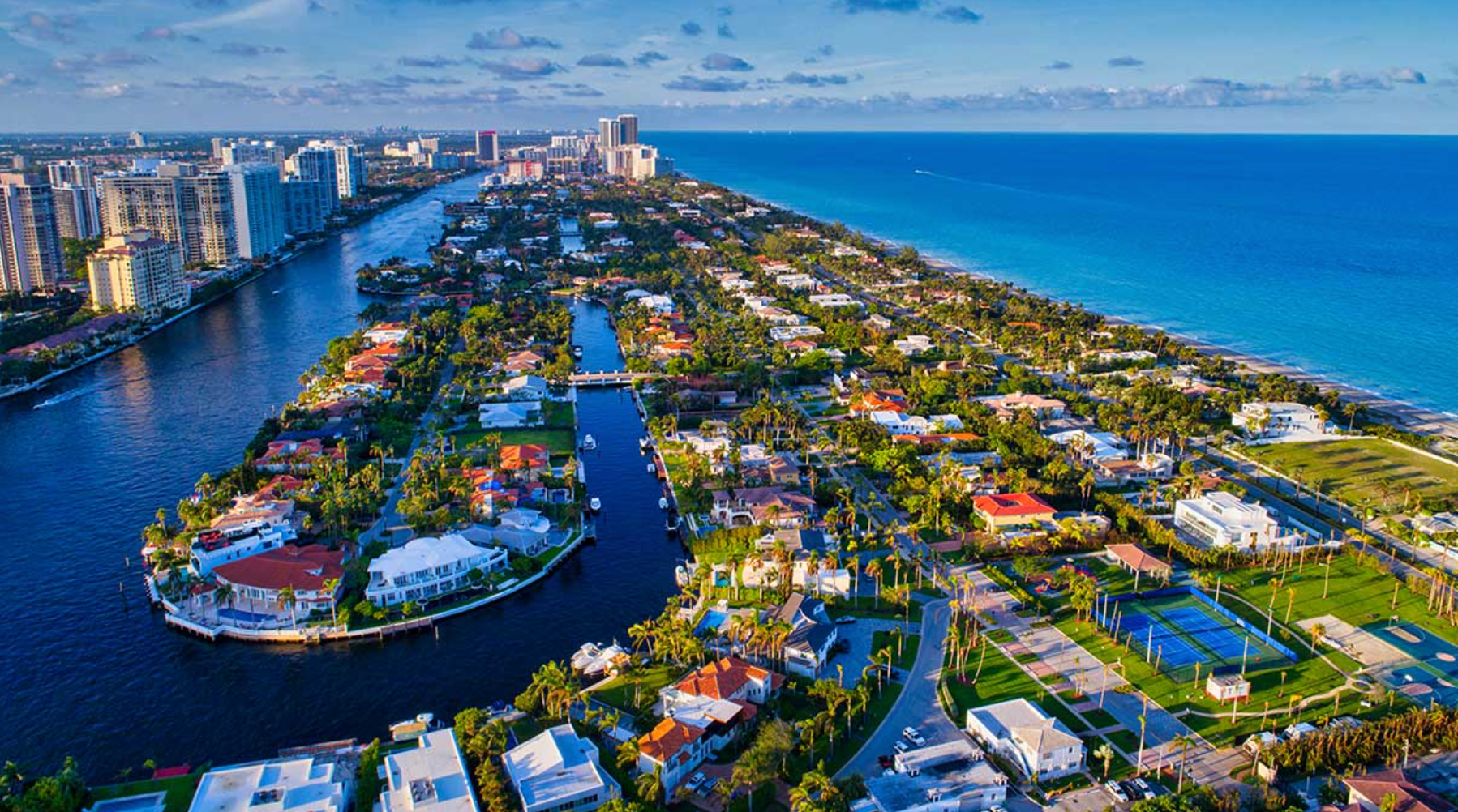 Aerial view of a coastal neighborhood with waterfront houses, canals, and a beach along the blue ocean.