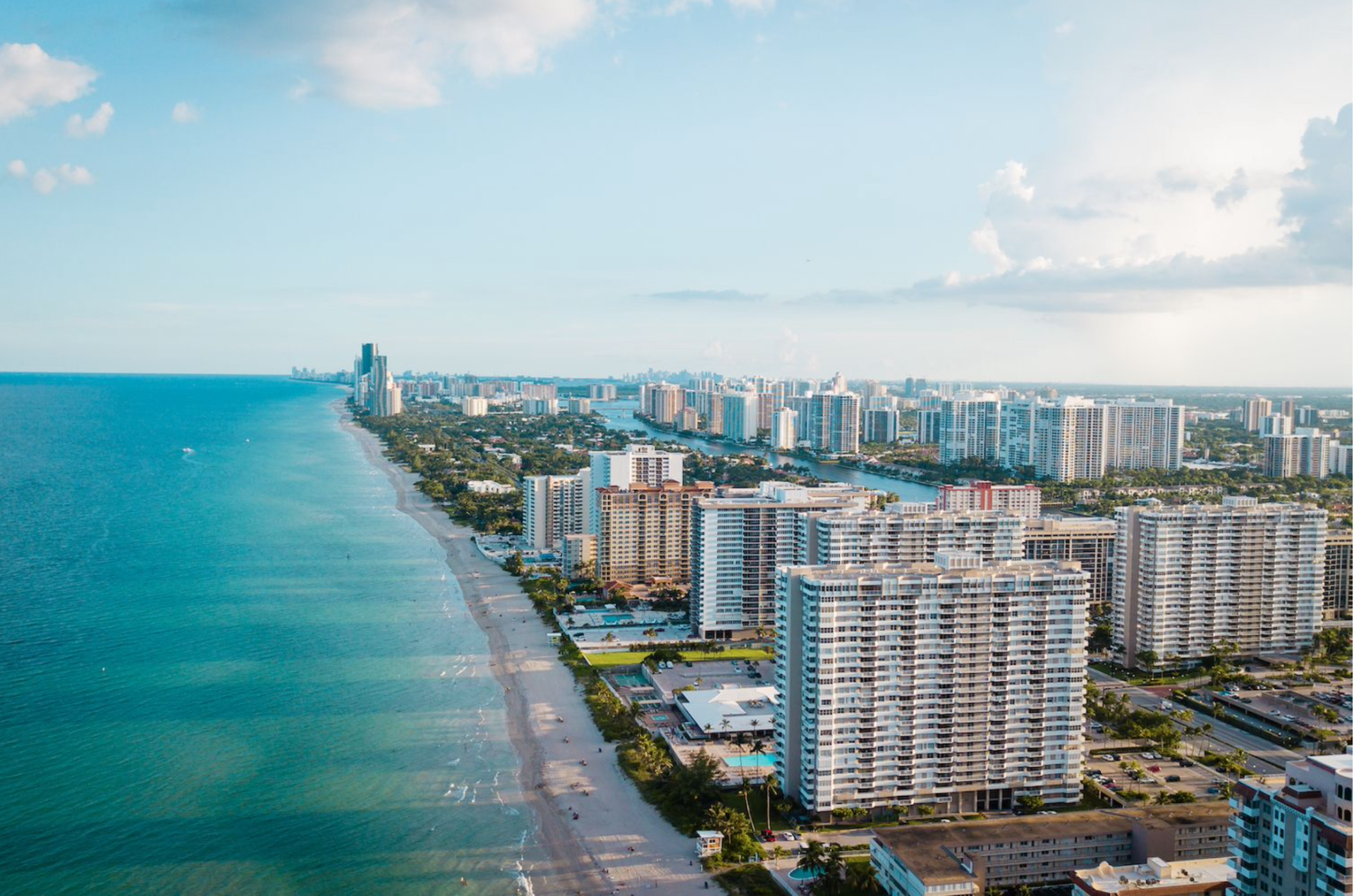 High-angle view of a sunny Florida coastline with high-rise apartment buildings bordering a wide, sandy beach.