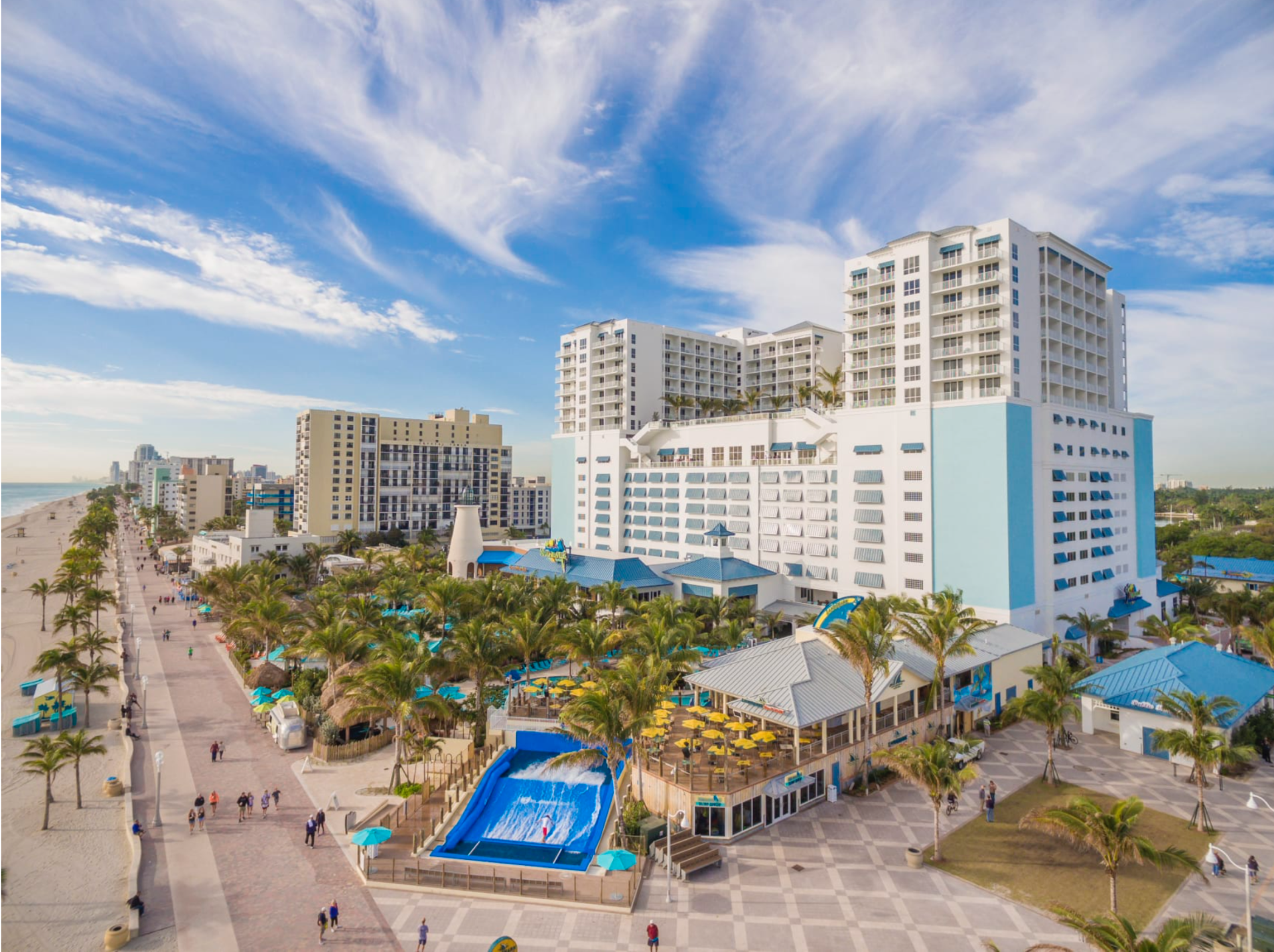 An elevated view of a beachfront resort with a blue wave simulator pool, palm trees, outdoor dining, and high-rise hotel.
